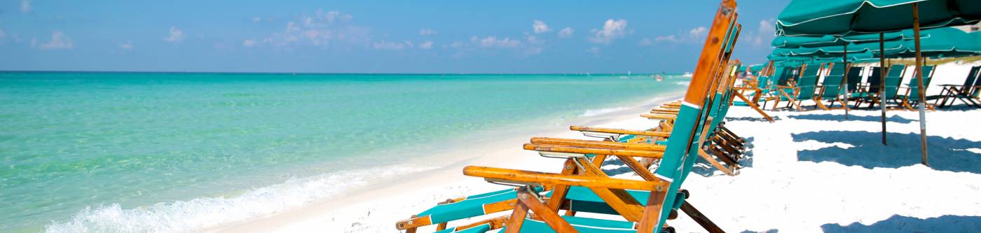 Wooden beach chairs with teal cushioning on a white sandy beach along green waters.